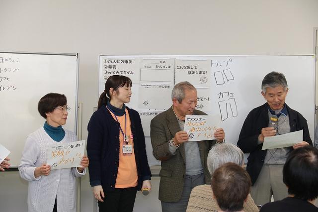 アイデアが貼り出されたホワイトボードを背に、中高齢の男女がマイクとアイデアが書かれた紙を手に持ち発表をしている写真