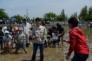 田植え中の子どもたちの前で赤いパーカーの男性と話をしている朝倉あきさんの様子の写真