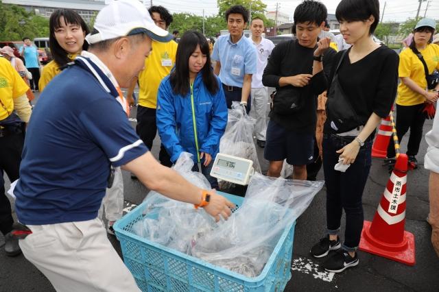 青い大きなケースに詰め込まれたゴミ入りの袋複数の重さを計量する男性と、それを見ている参加者の写真