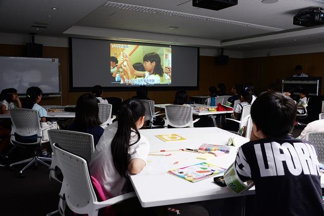 グループごとに分かれた机に座って、部屋前方のスクリーンに映し出された映像を見ている子ども達の写真