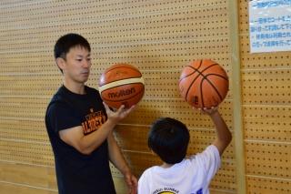 屋内の壁の側で、バスケットボールを持つ男の子に、ボールを持ちながら話しかけている男性の写真