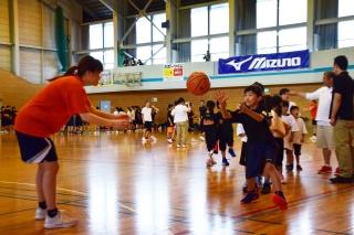 体育館内でバスケットボールをパスする女性と、それを受け取ろうとしている女子児童の写真
