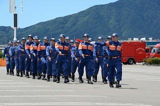 旗を持った男性を背に、青とオレンジの制服を着た消防士たちが、右を向いて行進している写真