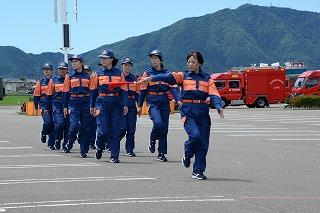旗を持った女性を背に、青とオレンジの制服を着た消防士たちが、右を向いて行進している写真
