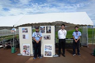 三人のスタッフが、資料写真の掲示されたボードとともに、テントの下に並んで立っている写真