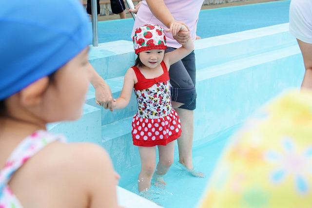 赤と白の水着を着た女の子が、両手をつながれてプールの中に入っていく写真