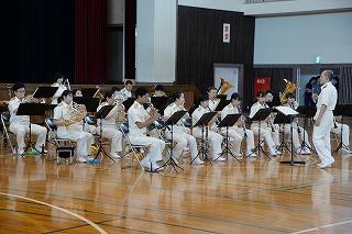 白い制服を着た人たちが、指揮者に向かって着席し、様々な楽器を演奏している写真