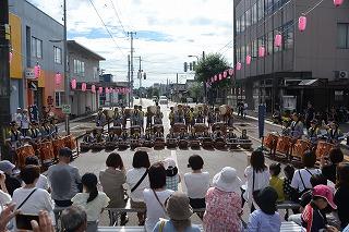道路を横切るように並び、和太鼓を演奏している子供たちと、それを見ている人たちの写真