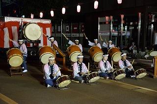 白いはっぴを着た人たちが、前列では座り、後列では立って、和太鼓を演奏している写真