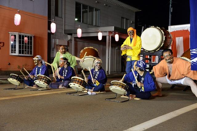 茶色いお面を付けた人たちが、道路に座って並び、和太鼓を演奏している写真