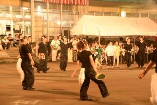黒い衣装に白い尻尾とお面を付けた人たちが、円形に並んで踊っている写真