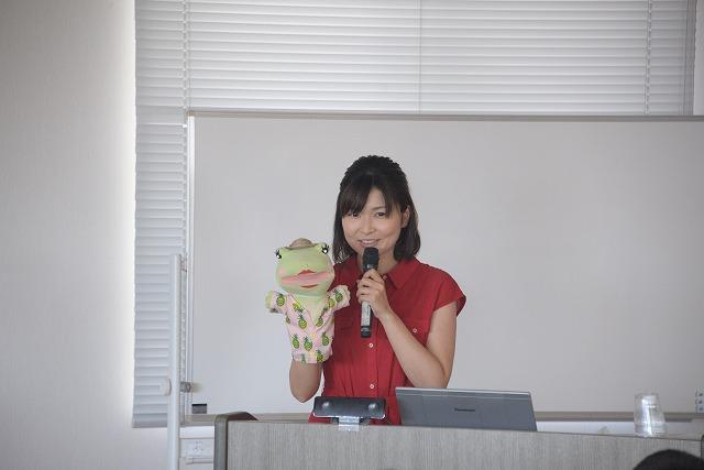 カエルの人形を手に講話する女性の写真