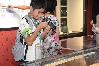 腕章をつけた児童がカメラを構えて展示物を撮影している写真