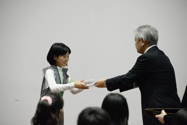 私服の女子生徒が、白髪で黒いスーツを着た男性から笑顔で贈り物を受け取っている写真