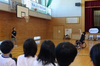 体育館で子ども達の見ている中、バスケットゴールへスリーポイントシュートを放つ車椅子に乗った男性の写真