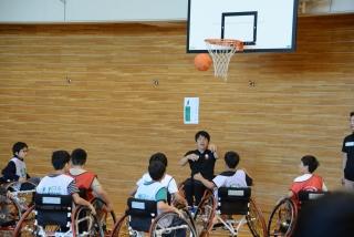バスケットゴールの周りに集っている車椅子に乗った生徒達と、車椅子に乗りながらバスケットボールを投げている男性の写真