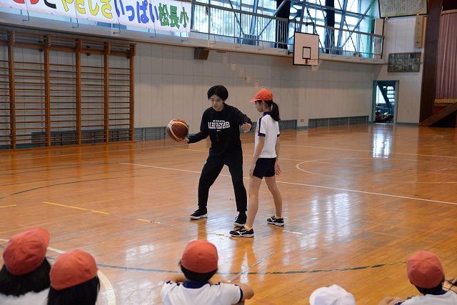 体育館内の床に座る子ども達の前で、バスケットボールの指導を身振りを加えながら説明している女性とそれに協力する女子生徒の写真