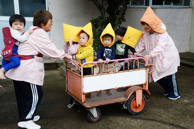 背に園児を背負いながら、防災頭巾を被った4人の園児を乗せた台車を運ぶ、2人の保育士の写真
