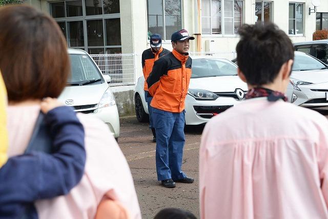 駐車場に集まった保育士と園児達を前に立って話をしている消防署の職員の写真