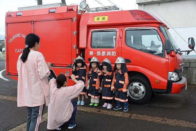 真っ赤な消防車を背に、消防服を着て並んだ園児達4人をスマホで撮影している保育士の写真