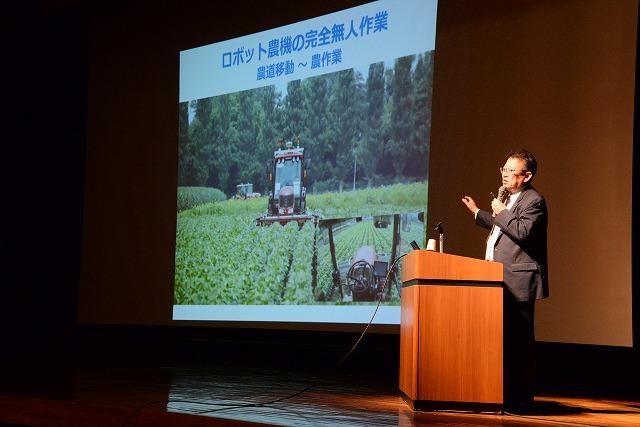 「ロボット農機の完全無人作業」と書かれたスライドと演壇で話すスーツ姿の教授の写真
