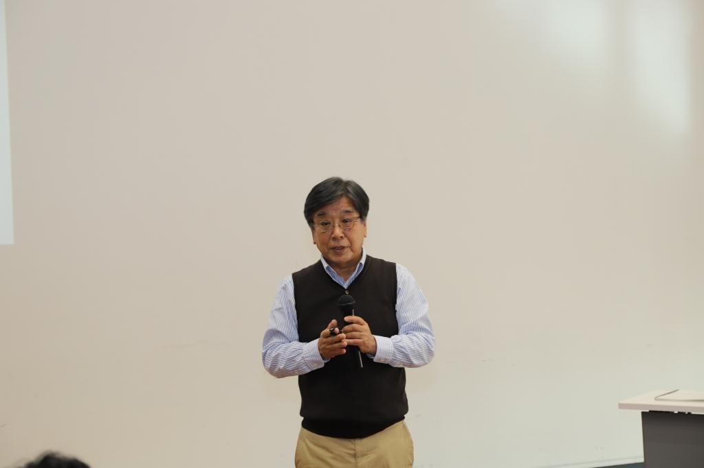 シャツとベストを着た志賀誠治さんがマイクを持ち参加者に向けて話している写真