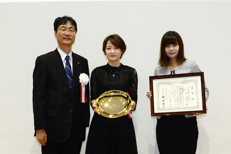 3人が並んでいて、向かって右側2人が女性で真ん中の女性が楯、右側の女性が賞状を持って表彰されている写真