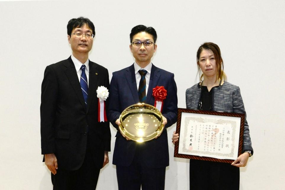 真ん中の男性が楯を持ち、右側の女性が賞状を持って3人で並んで表彰されている写真