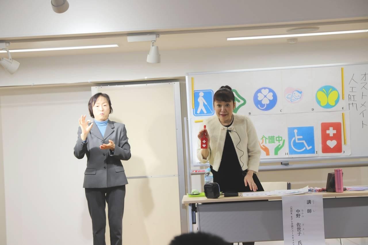 ヘルプマークを手にして話をしている講師の女性とその横で手話通訳をしている女性の様子の写真