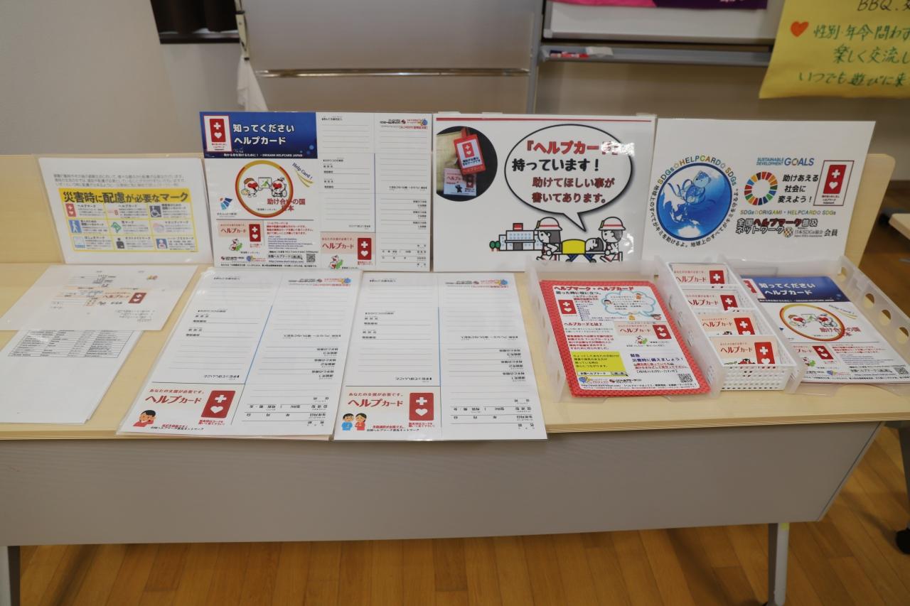 白い机の上に並べられたたくさんのヘルプカードと4つのヘルプマークの認知に関する説明が書かれた掲示物の写真