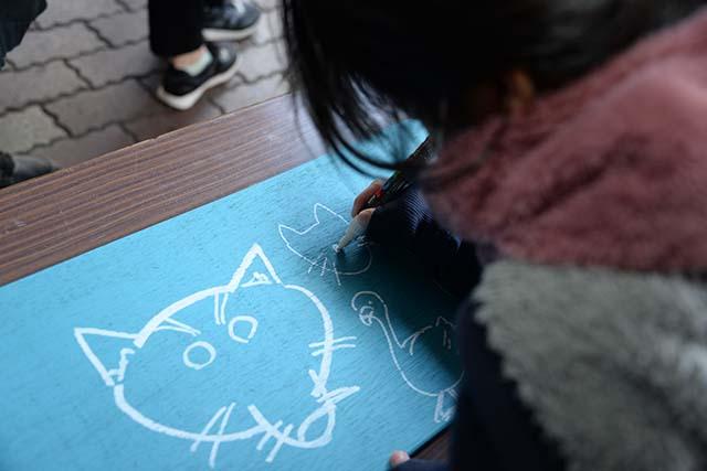 長机の上に敷かれている青い画用紙の上に白いペンで猫などを書いている女の子の様子の写真