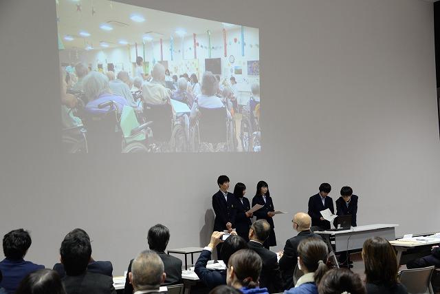 スクリーンを背に立ち発表をしている5人の学生達と、それを聞いている参加者達の写真