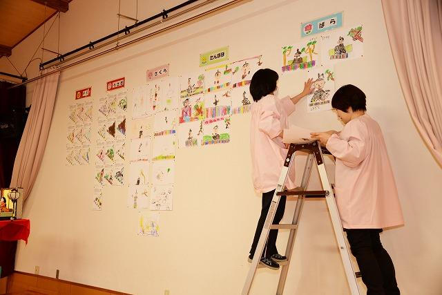 白い壁の一角に、脚立を使って子ども達が描いた絵、塗った絵を貼り付けている女性二人の写真