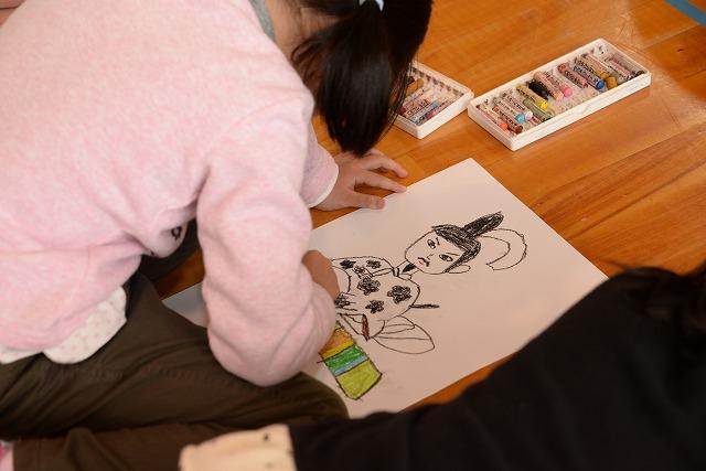 床に置いた白い紙にクレヨンで菅原道真の絵を描いている女の子の写真