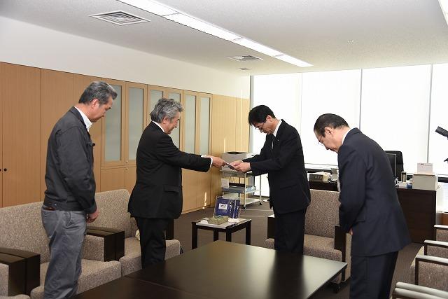 執務室で、封筒を手渡している男性と、頭を下げて受け取っている市長と職員の写真