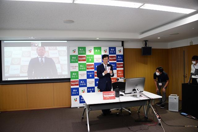 男性の写っているスクリーンの隣で、燕市のボードの前に立ち、封筒を持って見せている市長の写真