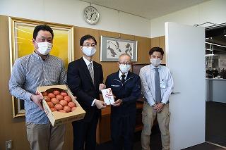 見やすいように掲げられたトマトの詰められた段ボール箱とのし袋を手に並ぶマスクを付けた4人の男性たちの写真
