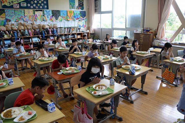 教室内に広げられた机と椅子に座って、盛り付けられた給食皿を前にいただきますのあいさつをしている子供たちの写真