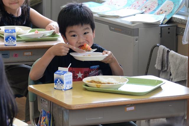 机に座って給食のトマトをスプーンで勢いよくかきこんで食べている紺色のシャツを着た男の子の写真
