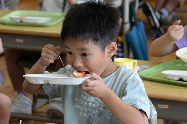 給食のトマトをスプーンで勢いよくかきこんで食べている灰色のシャツを着た男の子の写真
