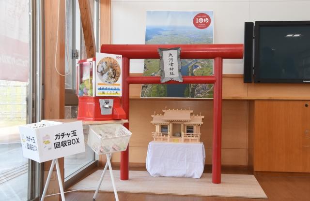 室内に置かれた白木造りの小型の神社と赤い鳥居、その脇に置かれたお守りガチャガチャ台の写真