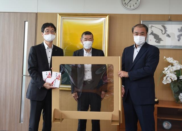 スーツ姿にマスクを付けた3人の男性たちがパーテーションを掲げながら笑顔で並んでいる写真