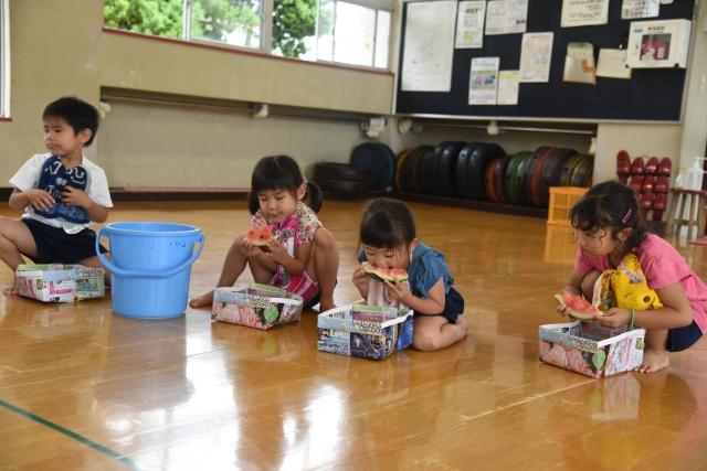 幼稚園の室内で種受けの手作りの紙皿を下にして、スイカにかぶりつく4人の子どもたちの写真