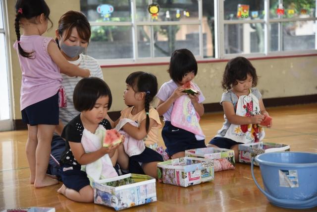 幼稚園の室内で種受けの手作りの紙皿を下にして、スイカを食べている5人の女の子たちと、後ろでそれを見守る女性の先生の写真