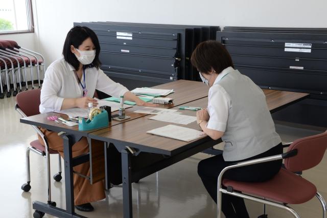 机を挟んで座り、並べられた用紙に向かっている女性と用紙に手をかざして説明する女性職員の写真