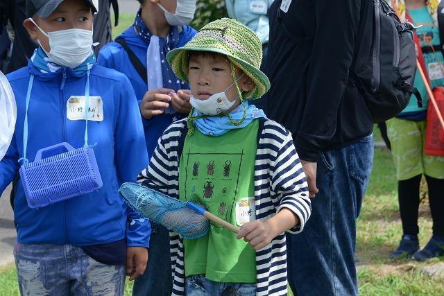 帽子を被りマスクを付け虫取り網を手に持ち、カブトムシやクワガタのイラストが描かれたTシャツを着た男の子の写真