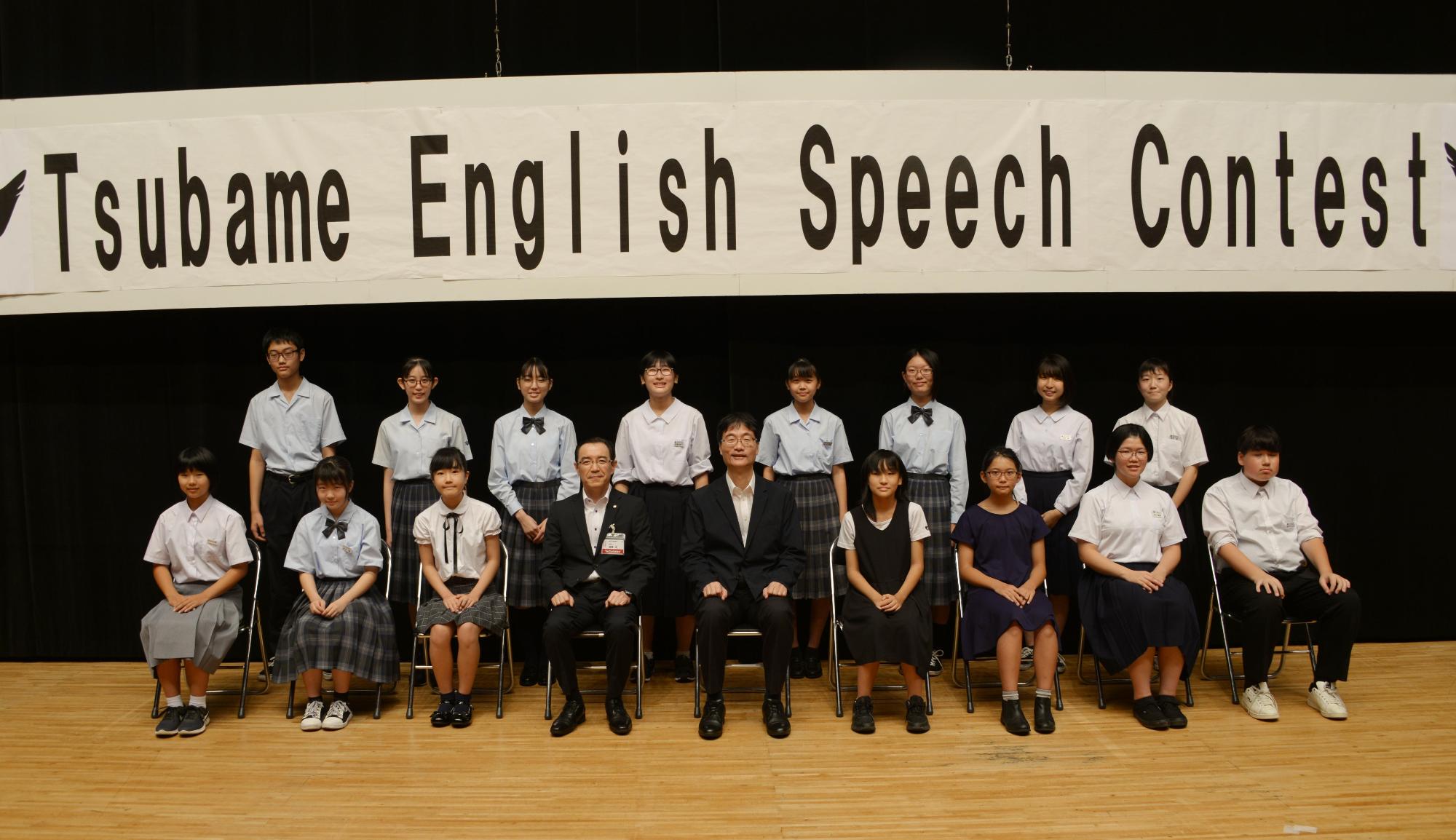 黒の暗幕の前に吊された「Tsubame English Speech Contest」と書かれた白い横断幕の下で、真ん中に座ったスーツ姿の2人の男性と共に記念撮影を行う男女の生徒たちの写真
