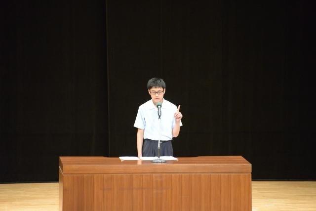 黒の暗幕を背にした壇上で、左手の人差し指を立てながらスピーチをする男子生徒の写真