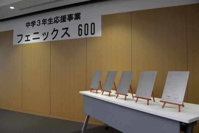 室内の壁際にかかる「中学三年生応援事業 フェニックス600」と書かれた白い垂れ幕と、その隣の机の上に立てられた5つの記念プレートの写真