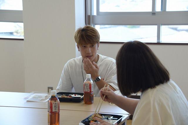 机の角を差し挟むように座りお弁当を食べながら談笑をする男性と女性の写真